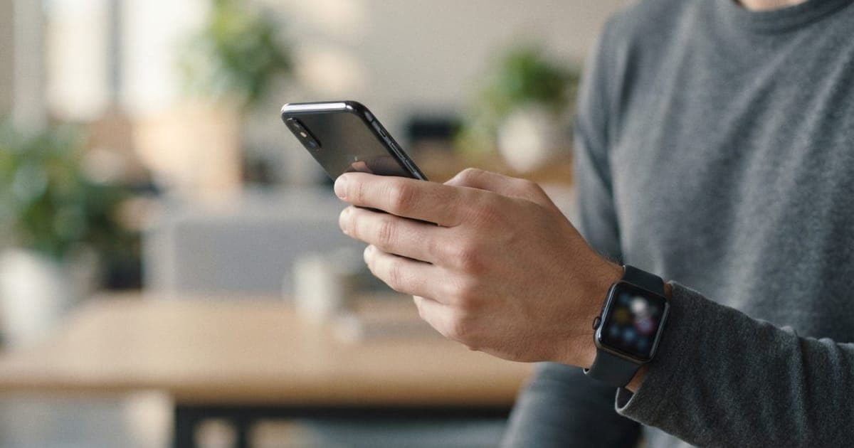 Close-up de mão segurando iPhone e Apple Watch com fundo neutro representando dispositivos do ecossistema Apple.