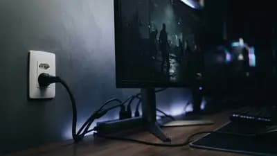 Close em monitor gamer com luzes RGB acesas, mostrando cabos de energia conectados a uma tomada próxima